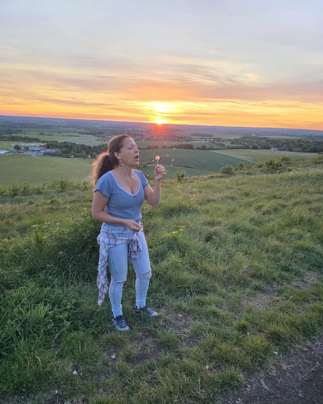 Valeria Cesario blowing a dandelion clock on a grassy hilltop at sunset